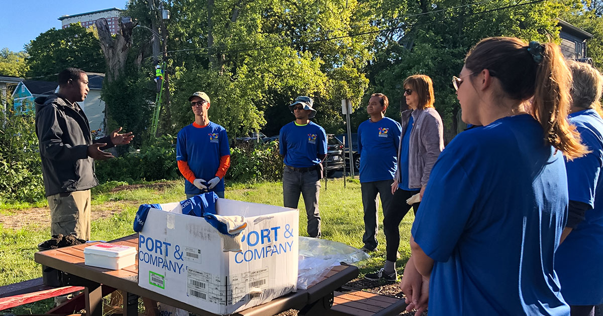 Group of people wearing blue Commitment to Community shirts, receiving instruction from man at Brick Gardens.