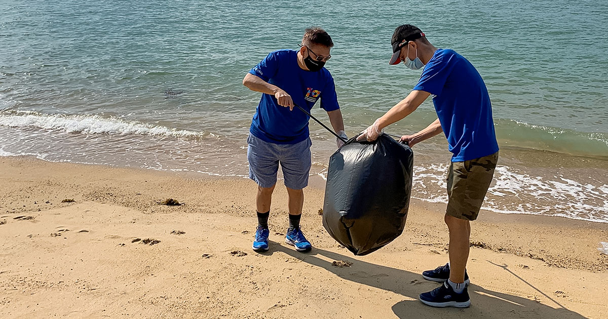 2 men wearing face masks and blue Commitment to Community shirts, cleaning trash on beach.